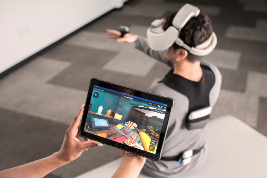 A physical therapist holds a tablet showing REAL System's tablet view while a client is wearing the VR headset while doing exercises.