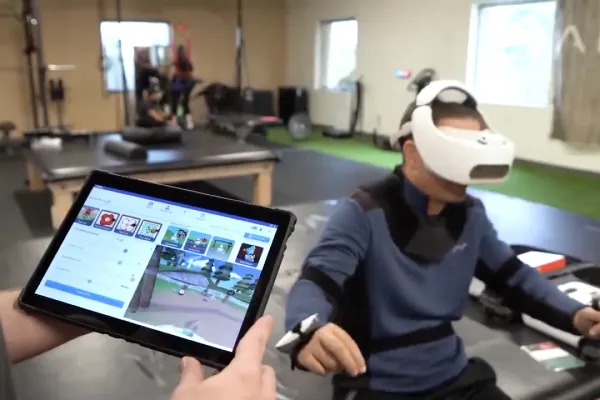 A physical therapist holds a tablet showing REAL System's tablet view while a client is wearing the VR headset while doing exercises.